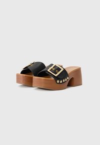 Mules pretos com plataforma e uma fita larga com uma fivela dourada, fixados a um salto em bloco de madeira castanha com detalhes de tachas.
