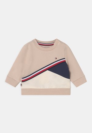 Sweat-shirt beige avec un design diagonal comportant des accents bleu marine, blanc et rouge. Tissu doux, col rond, fermeture à pression sur l'épaule.