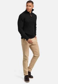 Schwarzer Strickpullover mit hohem Kragen und Reißverschluss, kombiniert mit beigen Hosen und braunen Schnürstiefeln, die ein strukturiertes Muster aufweisen.