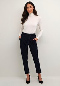 Suéter blanco de cuello alto combinado con pantalones sastre azul marino con cintura anudada. Zapatos de tacón altos negros. Textura de tela suave.