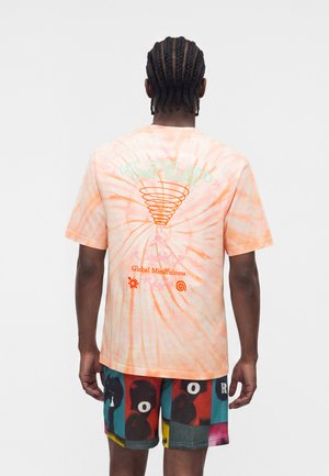 Fonott hajú férfi, aki narancssárga, spirál mintás, batikolt pólót és színes mintás rövidnadrágot visel, háttal áll.