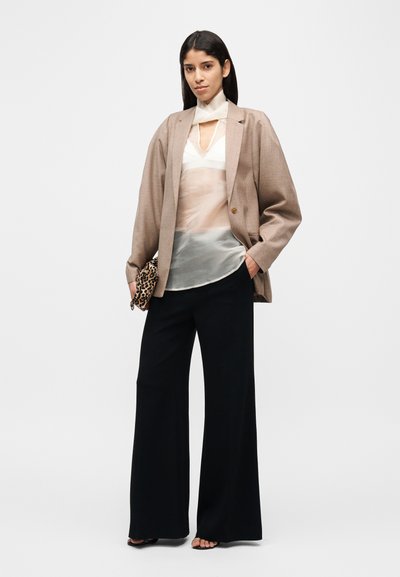 Blazer oversize beige, top blanc transparent avec un détail en forme de trou clé, pantalon noir à jambes larges, et pochette à motif. Esthétique minimaliste.