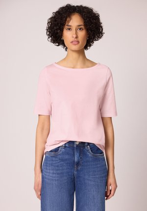 Femme aux cheveux courts et bouclés portant un t-shirt rose clair à manches courtes et un jean bleu, debout devant un fond uni.