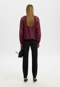 Viininpunainen läpikuultava pusero, jossa on liehukkeet hihnoissa, mustat räätälöidyt housut, kädessä pieni musta laukku, katsottuna takaa. Sileä materiaali, minimalistinen muotoilu.