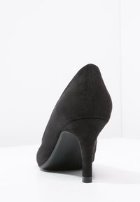 Scarpa nera in suede con tacco alto vista da dietro, caratterizzata da un sottile tacco a stiletto su una superficie bianca.