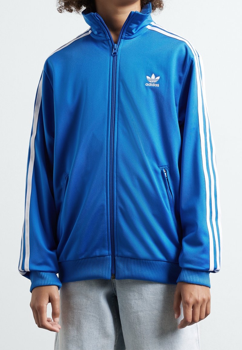 Modrá zip-up sportovní bunda vyrobená z hladkého polyesteru, s bílými pruhovanými detaily na rukávech a logem Adidas na hrudi.