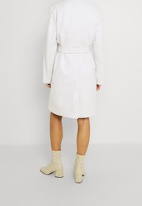 Jasnoszary płaszcz z paskiem o luźnym kroju, z długimi rękawami i rozcięciem z tyłu, zestawiony z beżowymi botkami za kostkę na kwadratowym obcasie.