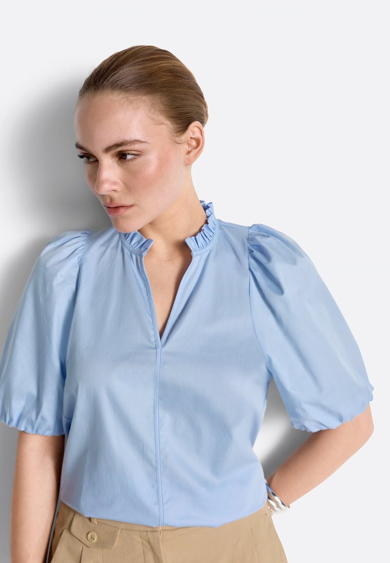 Femme aux cheveux tirés en arrière portant un chemisier bleu clair à manches bouffantes et col à volants, debout devant un fond blanc.