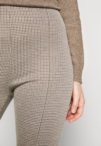 Hosen mit einem Hahnentrittmuster in Beige und Braun, mit einer taillierten Passform und weichem Material. Der Bund ist in der Nähe eines gestrickten Pullovers sichtbar.