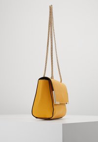 Žlutá kabelka s texturovaným povrchem, zlatým řetízkovým popruhem a obdélníkovou sponou. Má černý okraj podél horní části. Kompaktní, strukturovaný tvar.