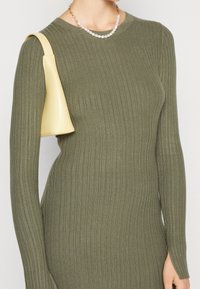 Robe tricotée côtelée vert olive avec des manches longues et une fente sur le côté, agrémentée d'un sac à main jaune pâle et d'un collier de perles.