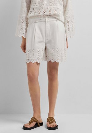 Witte shorts met hoge taille en geschulpte bloemige kanten zoom, gecombineerd met een bijpassende kanten blouse en bruine teenslippers op het model.