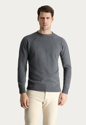 Maglione grigio lavorato a maglia con un design a trecce testurizzato sulle spalle, polsini e orlo a coste, indossato con pantaloni chiari.