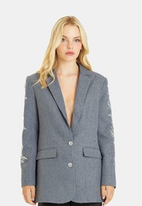 Blazer grigio con un design strutturato, scollatura a V profonda, ricami decorativi sulle maniche e due tasche frontali. Presenta una chiusura con due bottoni.