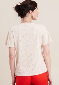 Kurzärmlige, cremefarbene Bluse mit einer Lochstickerei-Textur und diamantförmigen Mustern sowie einem gewellten Ausschnitt, kombiniert mit roten Hosen.