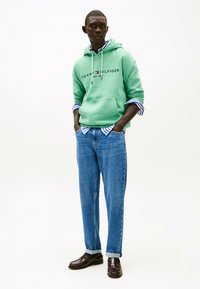 Sudadera verde con logo, superpuesta a una camisa de rayas azul y blanca, combinada con jeans azul claro y zapatos marrones oscuros.