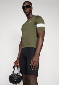 Tricou de ciclism verde măslin cu mâneci scurte, cu detalii albe pe braț, asortat cu pantaloni scurți din material căptușit negri. Modelul ține un casca neagră de bicicletă.