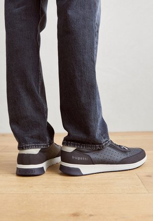 Herrskor från Bugatti i mörkgrått med perforerade detaljer, gummisula och beigefärgade accenter. Bärs tillsammans med mörka jeans på ett trägolv.