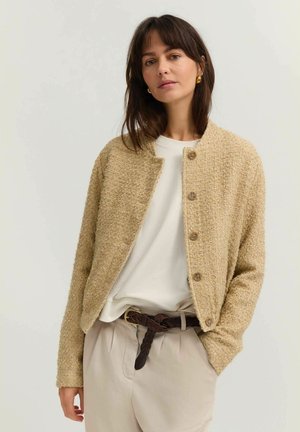 Frau trägt eine strukturierte beige, geknöpfte Strickjacke über einem weißen Hemd, beige Hose mit hoher Taille und einen geflochtenen dunkelbraunen Gürtel.