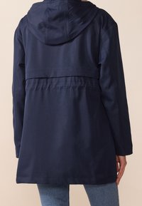 Marinblå vattentät jacka med huva, med en indragen midja, långa ärmar och en mjuk textur. Minimalistisk design utan synlig hårdvara.