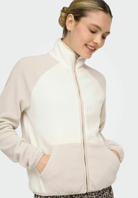 Chaqueta de forro polar en crema y beige, con cuello alto, cierre de cremallera, mangas ranglán y bolsillos frontales. Textura suave con elementos contrastantes.
