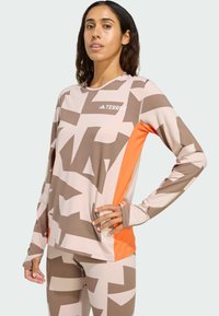 Beige atletische top met lange mouwen, bruine geometrische patronen en oranje zijpanelen. De stof oogt glad en aansluitend, met een ronde hals.