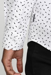 La mano de un hombre junto a la manga larga blanca de una camisa con pequeños patrones de bigotes negros y pantalones negros.