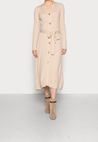 Abito a coste beige con maniche lunghe, scollo a V, chiusura con bottoni e cintura in vita. Abbinato a stivaletti beige alla caviglia con cinturini marroni.