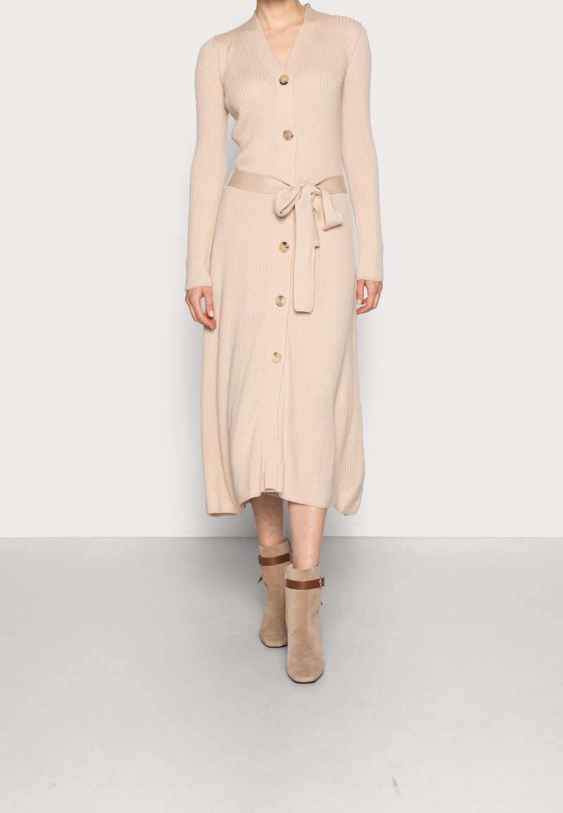 Abito a coste beige con maniche lunghe, scollo a V, chiusura con bottoni e cintura in vita. Abbinato a stivaletti beige alla caviglia con cinturini marroni.