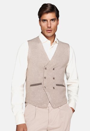 Man wearing a beige herringbone double-breasted vest over a white shirt and light beige trousers, standing with one hand in pocket.