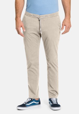 Uomo che indossa pantaloni beige chiaro con coulisse, camicia blu e sneakers blu e bianche, in piedi davanti a uno sfondo semplice.