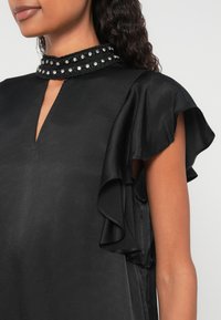 Čierna blúzka s výstrihom do tvaru V a volánovými rukávmi. Má zdobený golier so sequínmi. Hladká látka s jemnou textúrou.