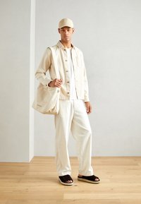 Giacca camicia beige, t-shirt bianca e pantaloni larghi, sandali scuri, cappellino abbinato e una borsa tote beige con dettagli ricamati.