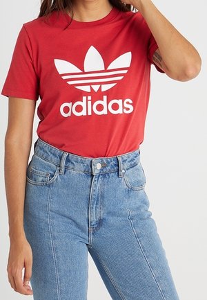 Kobieta nosząca czerwoną koszulkę Adidas z białym logo i niebieskie jeansy z wysokim stanem, stojąca na tle jednolitego jasnego tła.