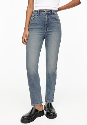 Jean bleu délavé taille haute associé à des chaussures noires sans lacets avec coutures blanches sur fond blanc.