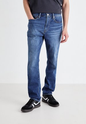 Blå denimjeans med rett benfitt, fem lommer og subtil falming. Kombinert med svarte joggesko med hvite detaljer.
