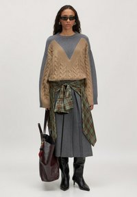 Vrouw in een beige en grijze kabelgebreide trui, grijze geplooide rok, een geruit shirt om de taille geknoopt, zwarte laarzen, zonnebril en met een grote donkere schoudertas in de hand.