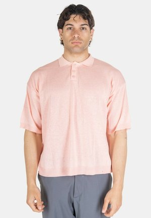 Uomo che indossa una polo a maniche corte in maglia rosa chiaro e pantaloni grigi, in piedi davanti a uno sfondo bianco semplice.