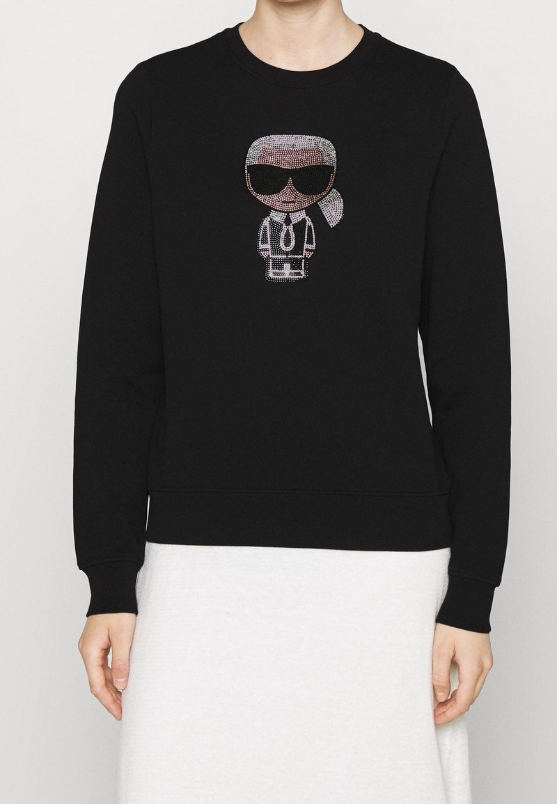 Sweat-shirt noir à manches longues avec un motif en strass représentant une personne stylisée avec des lunettes de soleil, des cheveux blancs et une cravate.