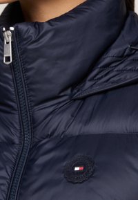 Marineblaue Daunenjacke mit hohem Kragen, ausgestattet mit einem Reißverschluss und einem runden Logo-Patch mit roten, weißen und blauen Akzenten. Glatte Textur.