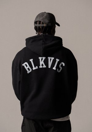 Personne aux cheveux tressés portant une casquette grise et un sweat à capuche noir avec "BLKVIS" imprimé dans le dos, debout devant un fond uni.