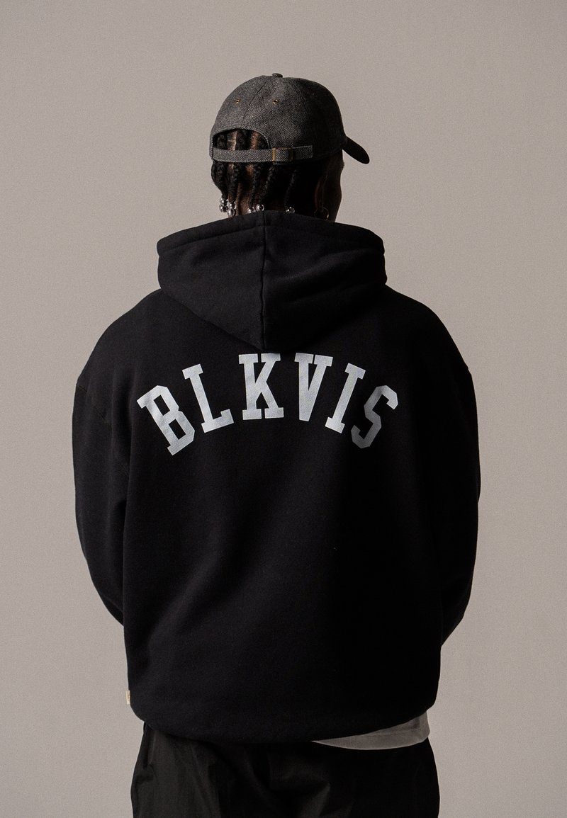 Personne aux cheveux tressés portant une casquette grise et un sweat à capuche noir avec "BLKVIS" imprimé dans le dos, debout devant un fond uni.