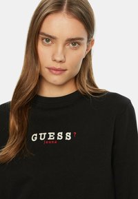 Felpa nera con colletto tondo, con ricamo della scritta "GUESS?" in bianco e rosso, e "jeans" in rosso. Texture liscia, design casual.