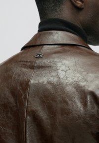 Bruine leren jas met een licht gerimpelde textuur, een nekkraag met inkeping en een zilveren logo-accent op de achterkant. Zwarte voering van de kraag zichtbaar.