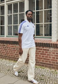 Polo shirt bianco con strisce blu, logo sul petto, abbinato a pantaloni larghi color crema e sneakers bianche con dettagli rossi.