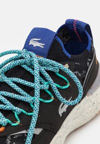 Zapatilla deportiva negra con un acabado texturizado, acentos en azul vibrante, cordones multicolores, suela moteada y un logotipo de Lacoste en el talón.