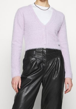 Personne portant un cardigan court duveteux lavande boutonné sur une chemise blanche, avec un pantalon en cuir noir taille haute et un collier chaîne en argent.