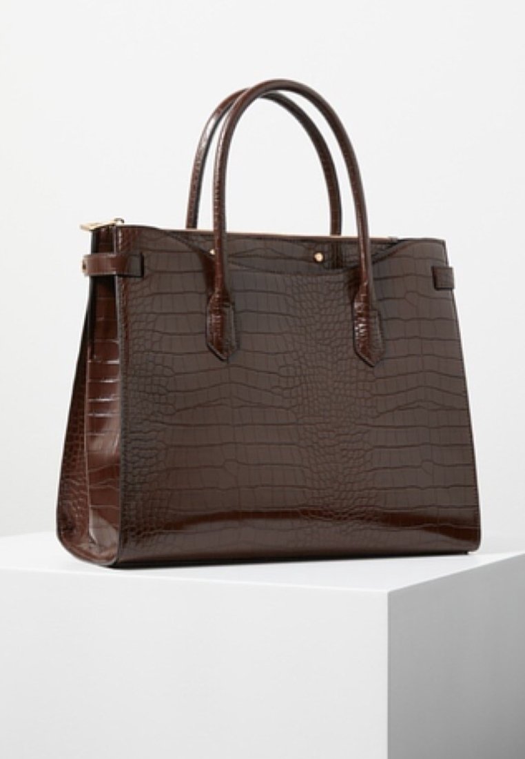 Sac à main en cuir marron foncé brillant avec motif crocodile, deux poignées arrondies et une forme rectangulaire structurée sur un piédestal blanc.