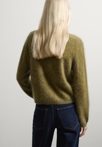 Suéter de color verde oliva, difuso, con un corte holgado y mangas largas, combinado con jeans de mezclilla azul oscuro con bolsillos traseros y corte recto.