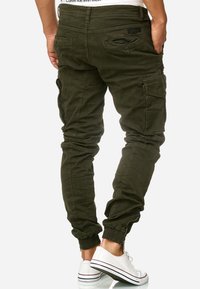 Hombre vistiendo pantalones cargo jogger en color verde olivo y zapatillas blancas de caña baja, de pie con las manos en los bolsillos, vista trasera.